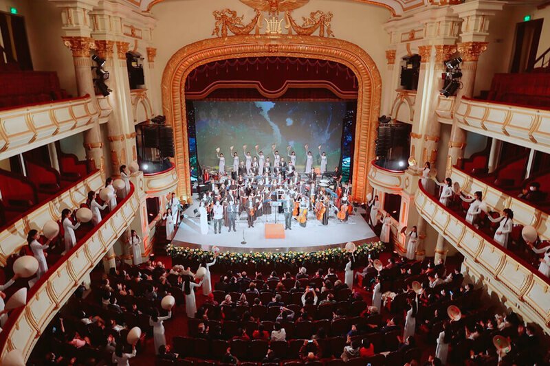 Salle de l'Opéra de Saïgon en plein concert, orchestre sur scène, public dans les balcons et le parterre, décoration rouge cerise et or