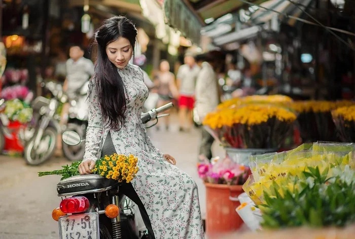 Jeune femme en áo dài garée à scooter dans une allée fleurie du marché de Ho Thi Ky