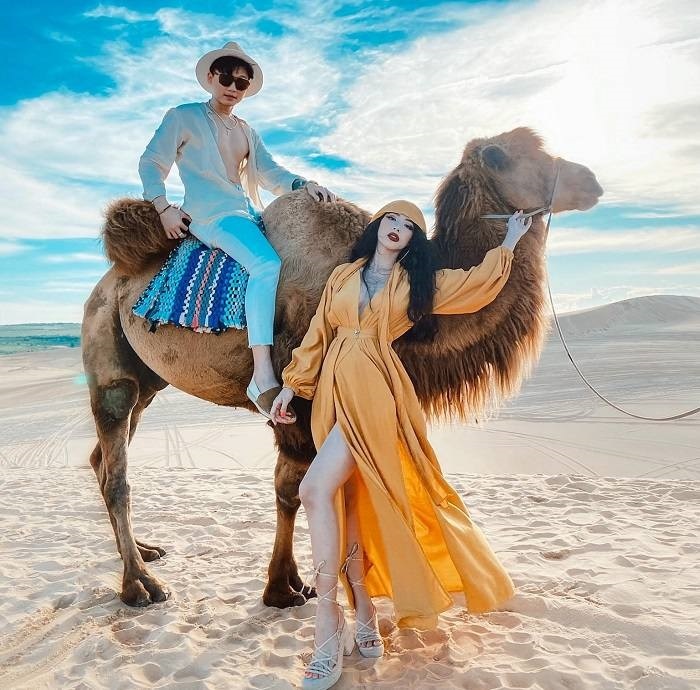 Trois jeunes femmes en robe orange posant avec un chameau sur les dunes blanches