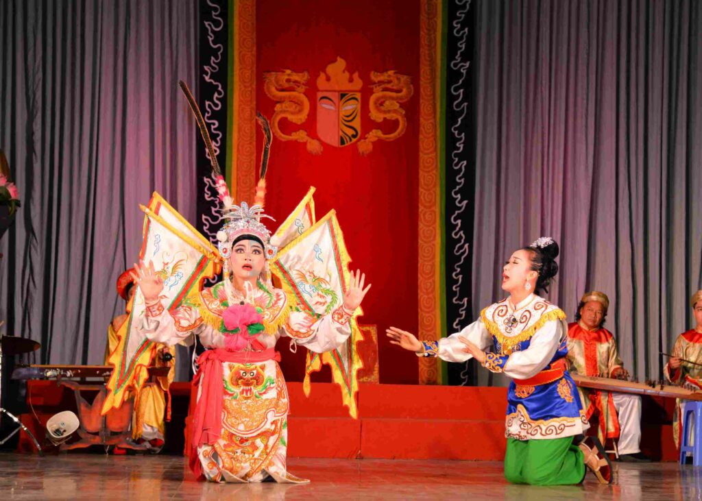 Artiste de Tuồng en costume élaboré avec drapeaux et musiciens en arrière-plan